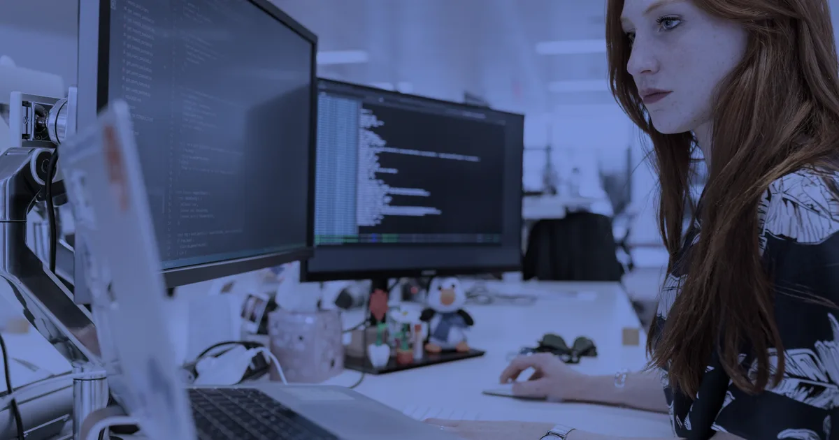 Software engineer reviewing code on two monitors in an open-plan office