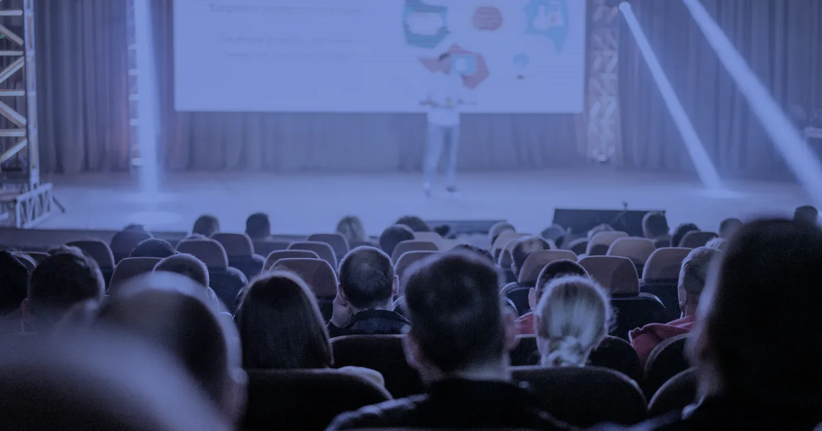 Audience at a conference watching a software vendor present a product demo on a large stage screen